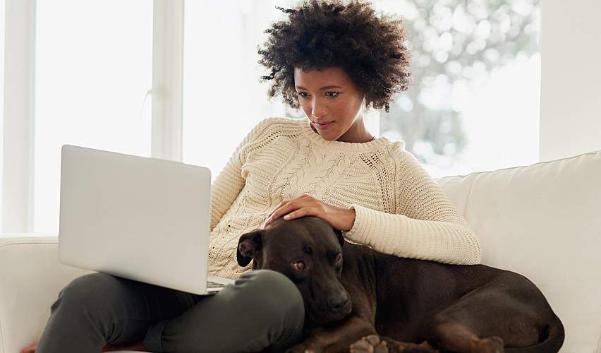 Woman uses computer while snuggling her dog on couch.