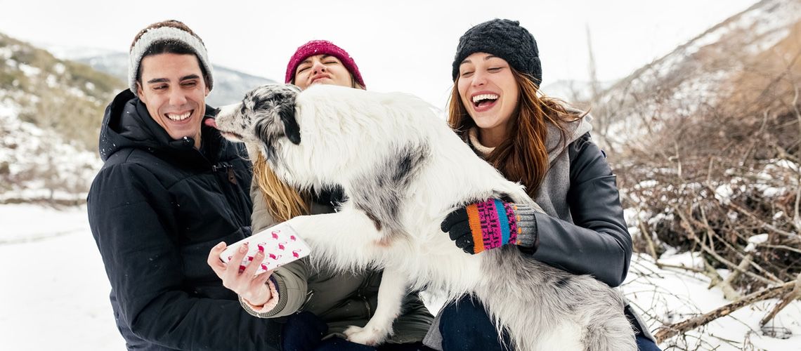 Group of young adults playing with a dog in the snow while taking a selfie.