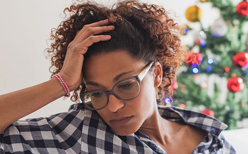A young woman wearing glasses appears stressed in front of a Christmas tree