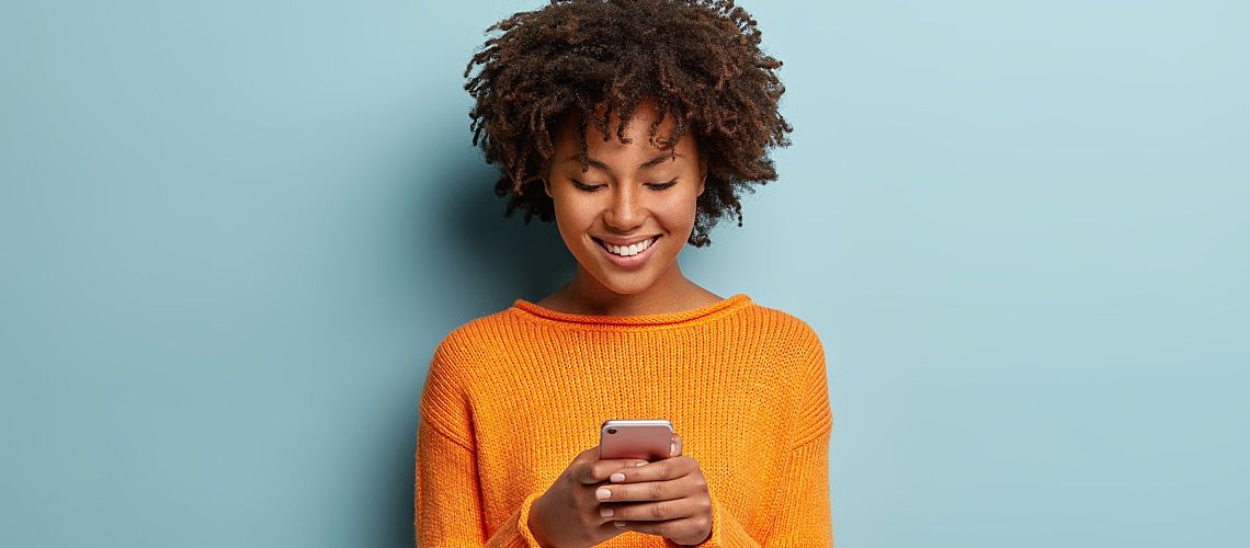 A young, pretty Black girl smiles while texting on a smartphone.