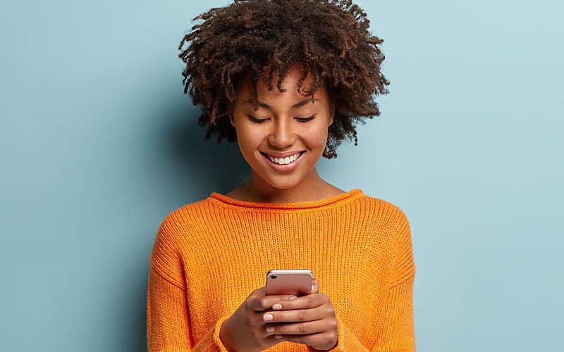 A young, pretty Black girl smiles while texting on a smartphone.
