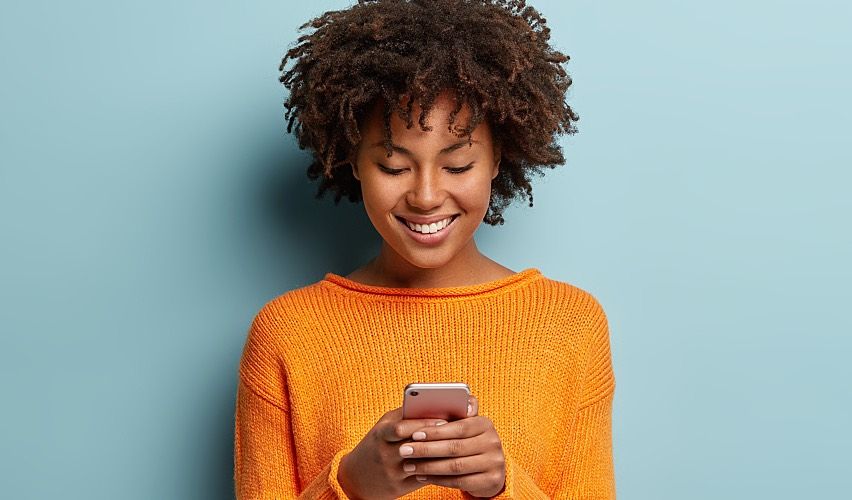 A young, pretty Black girl smiles while texting on a smartphone.