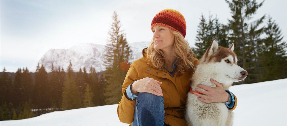 Woman sitting in the snow with her dog while looking off in the distance.