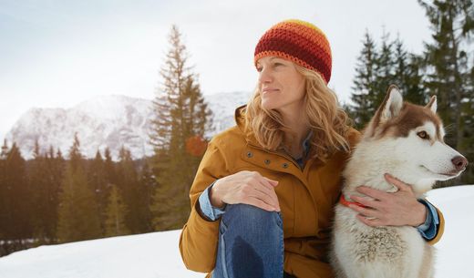 Woman sitting in the snow with her dog while looking off in the distance.