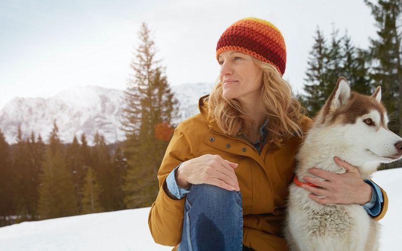 Woman sitting in the snow with her dog while looking off in the distance.